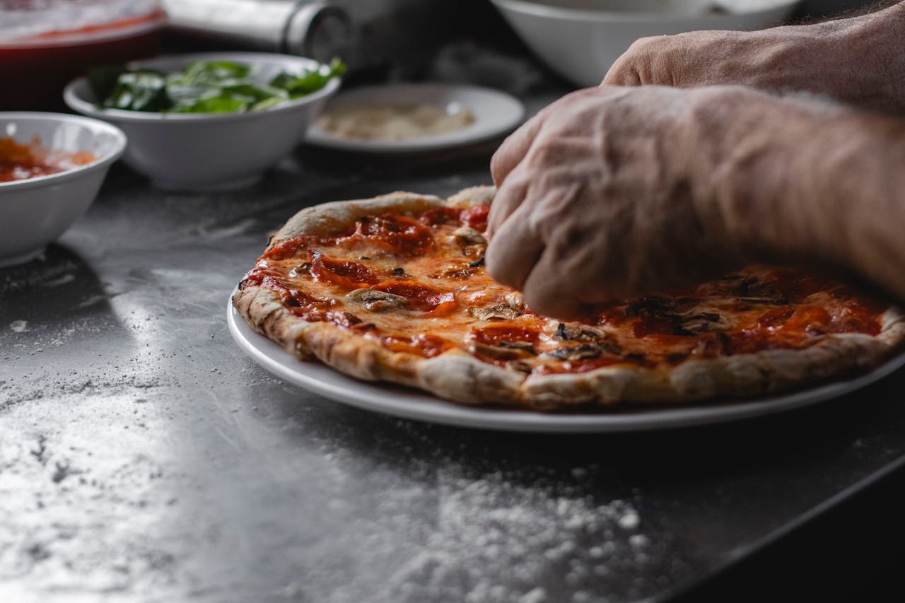 Close-up of mans hands preparing a freshly cooked pizza with toppings in the kitchen.
