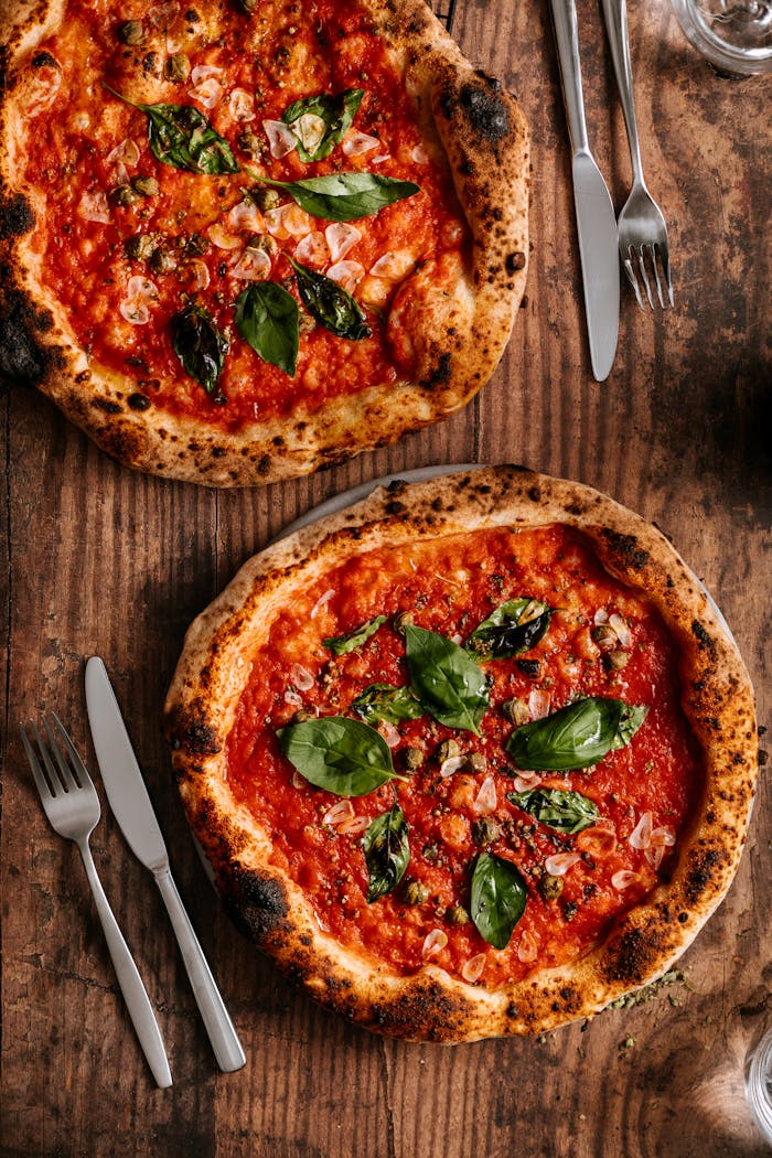 Top view of two Neapolitan pizzas with fresh ingredients and silverware on a rustic wooden table.